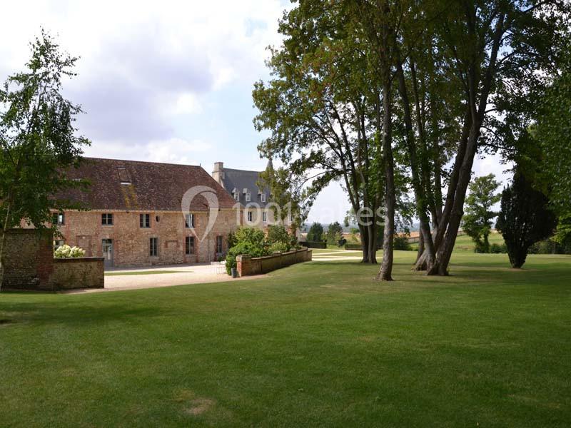 Bâtisse en pierre avec toit en ardoise entourée d'un grand jardin arboré sous un ciel partiellement nuageux.