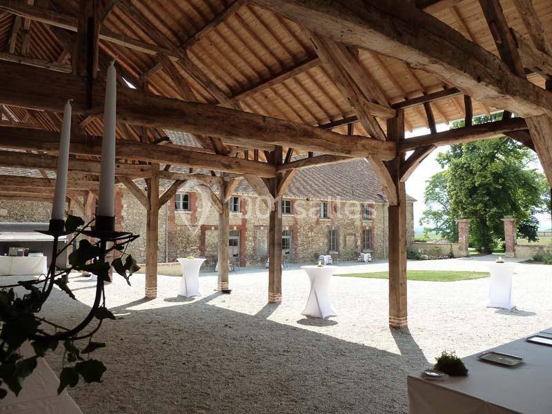Structure en bois d'une grange ouverte donnant sur une cour pavée avec des tables hautes et un bâtiment en pierre.