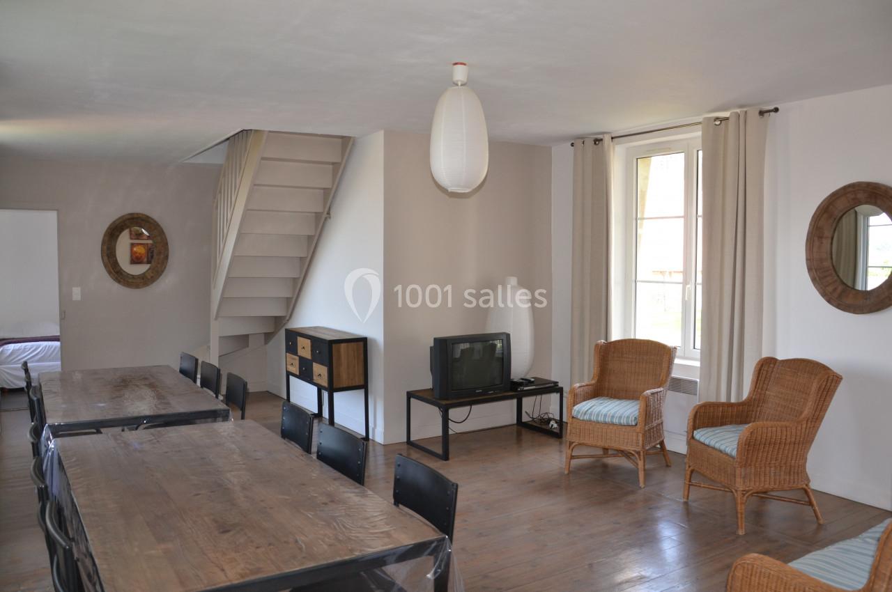 Salle de séjour lumineuse avec table en bois, chaises noires, fauteuils en rotin, télévision et escalier menant à l'étage.