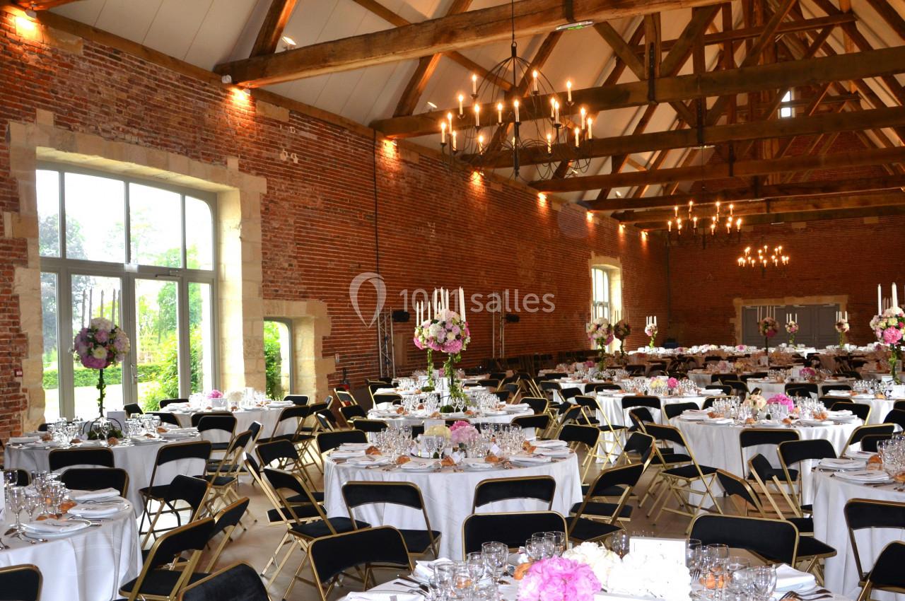 Salle de réception en briques avec poutres apparentes, tables rondes dressées et décorations florales élégantes.