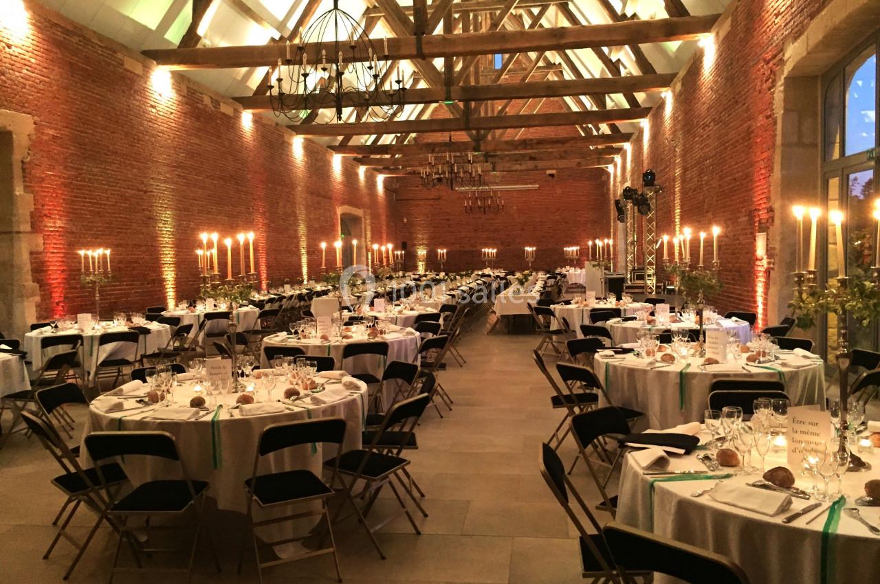 Salle de réception avec tables rondes dressées, chaises noires et éclairage chaleureux sous une charpente apparente.