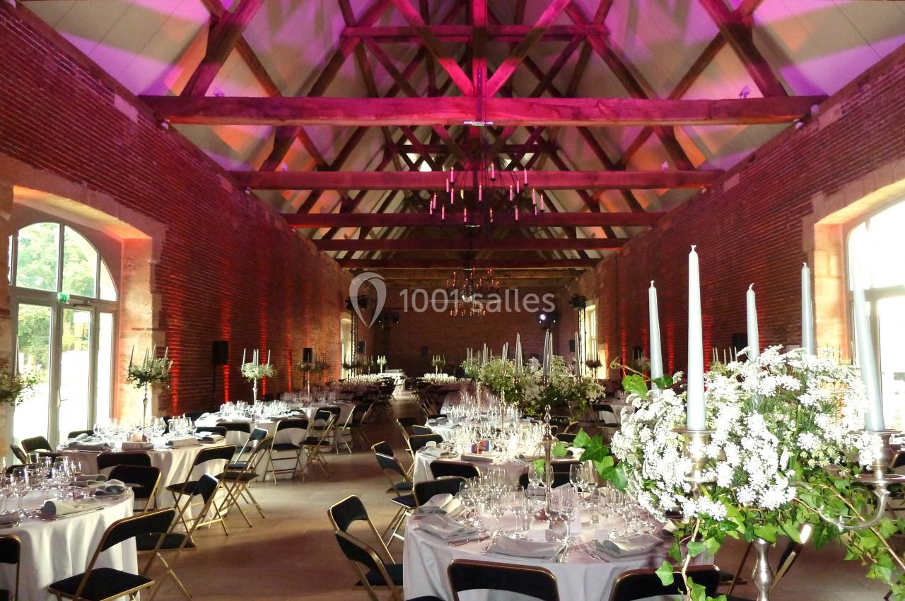 Grande salle de réception avec poutres apparentes, tables rondes dressées et décorations florales blanches.
