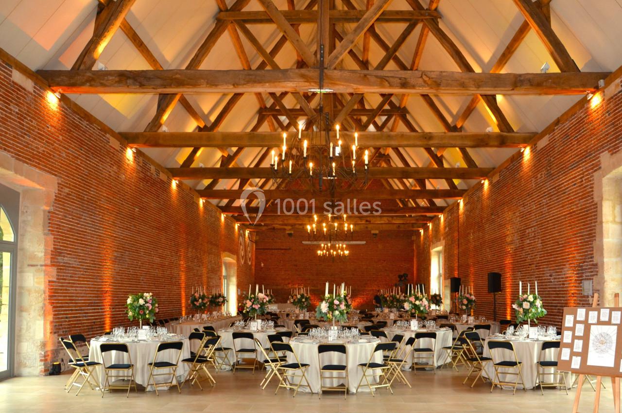 Salle de réception avec murs en briques, poutres apparentes, tables décorées et chandeliers suspendus.