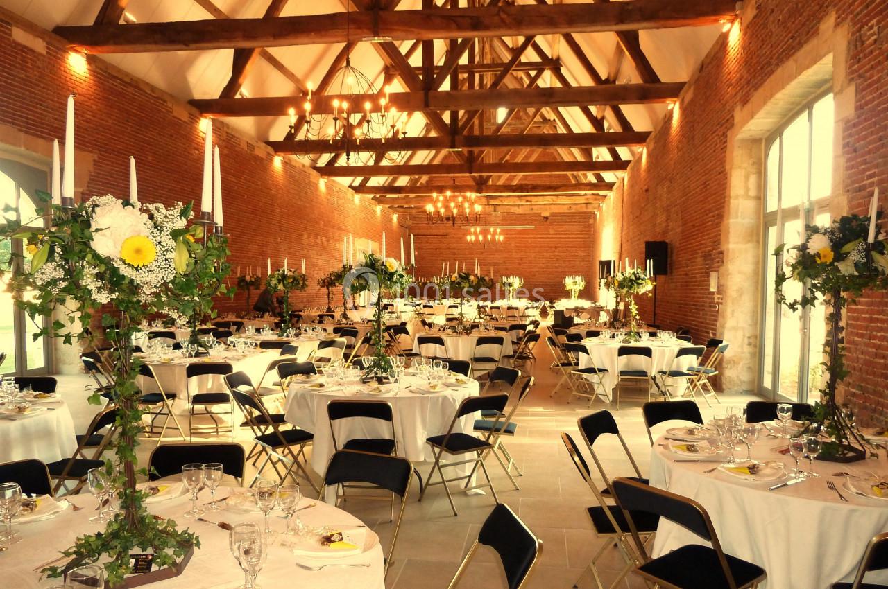 Salle de réception avec tables rondes dressées, décor floral et poutres apparentes sous un plafond haut.