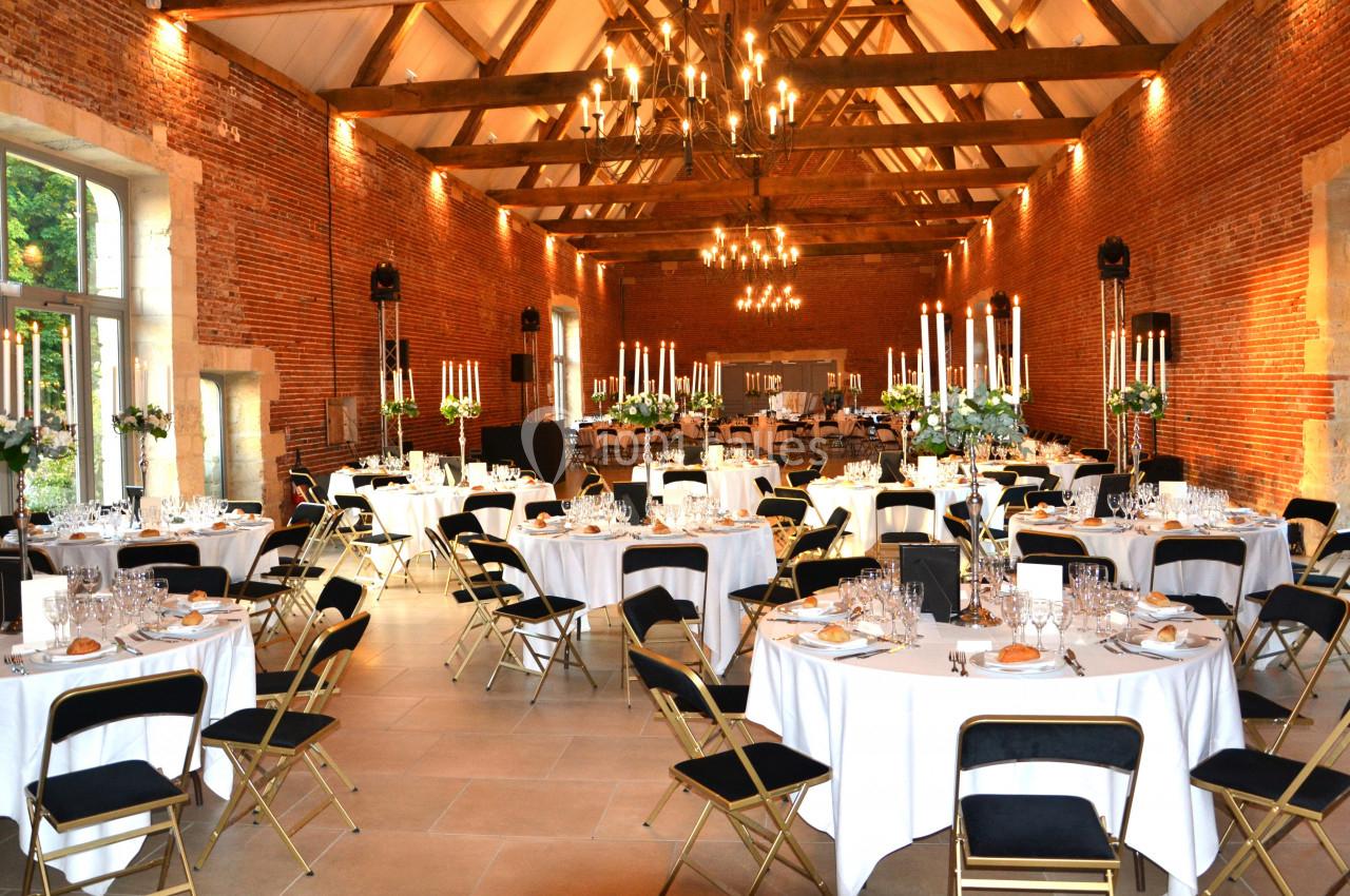 Salle de réception en briques rouges avec tables rondes dressées, chandeliers et grandes baies vitrées.
