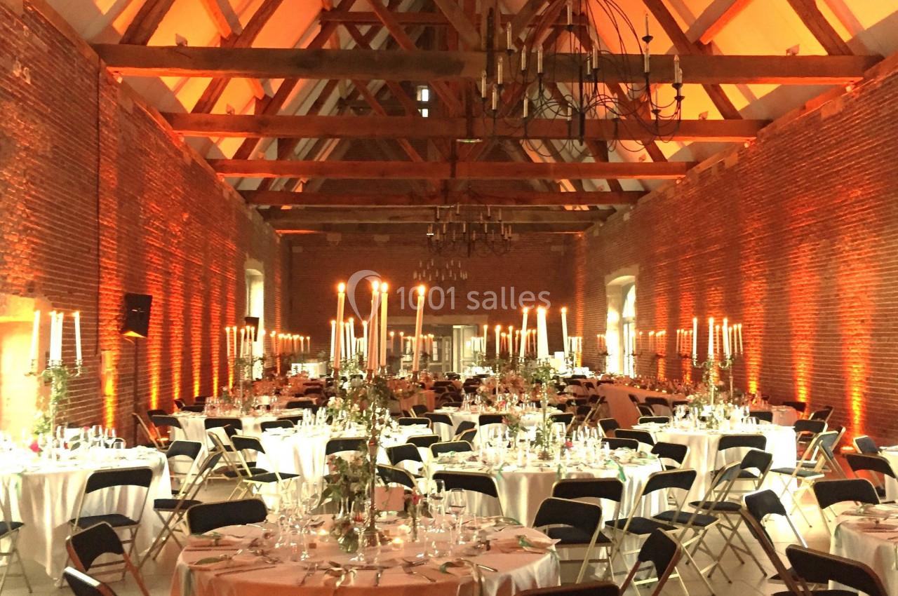Salle de réception avec murs en briques, poutres apparentes et tables rondes décorées de chandeliers et fleurs.