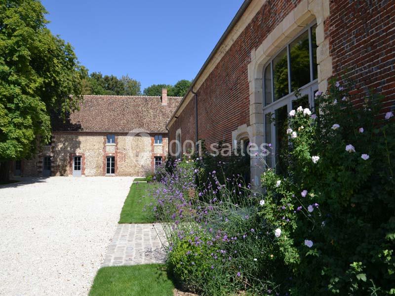 Cour pavée bordée de bâtiments en pierre et en briques, avec des fleurs et des arbres en arrière-plan.