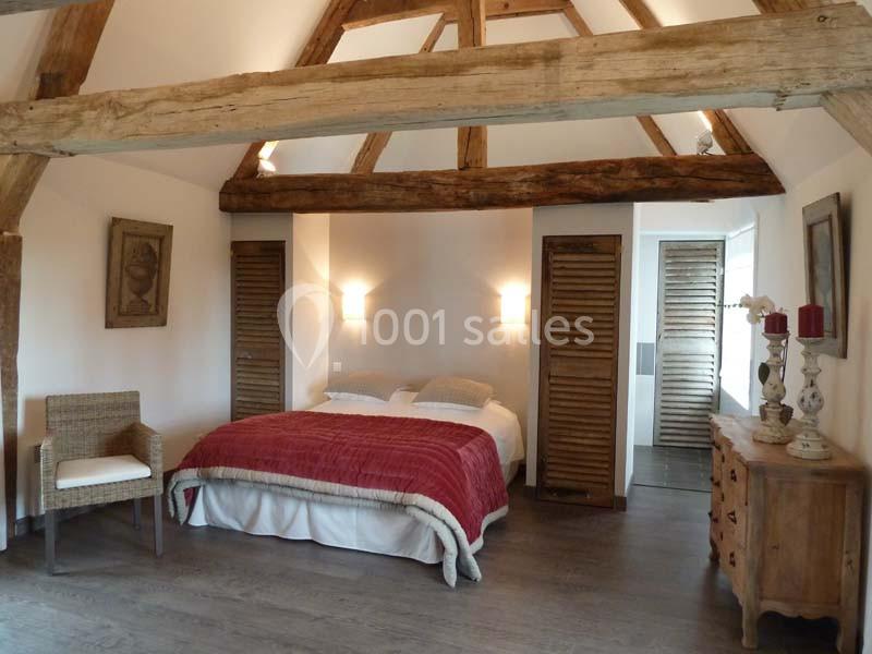 Chambre lumineuse avec poutres apparentes, lit double, mobilier en bois et chaise en osier sur un sol en parquet.