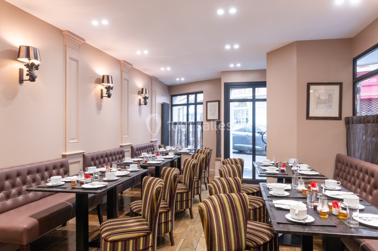Salle de restaurant lumineuse avec tables dressées, chaises rayées et murs beige, vue sur des fenêtres donnant sur la rue.