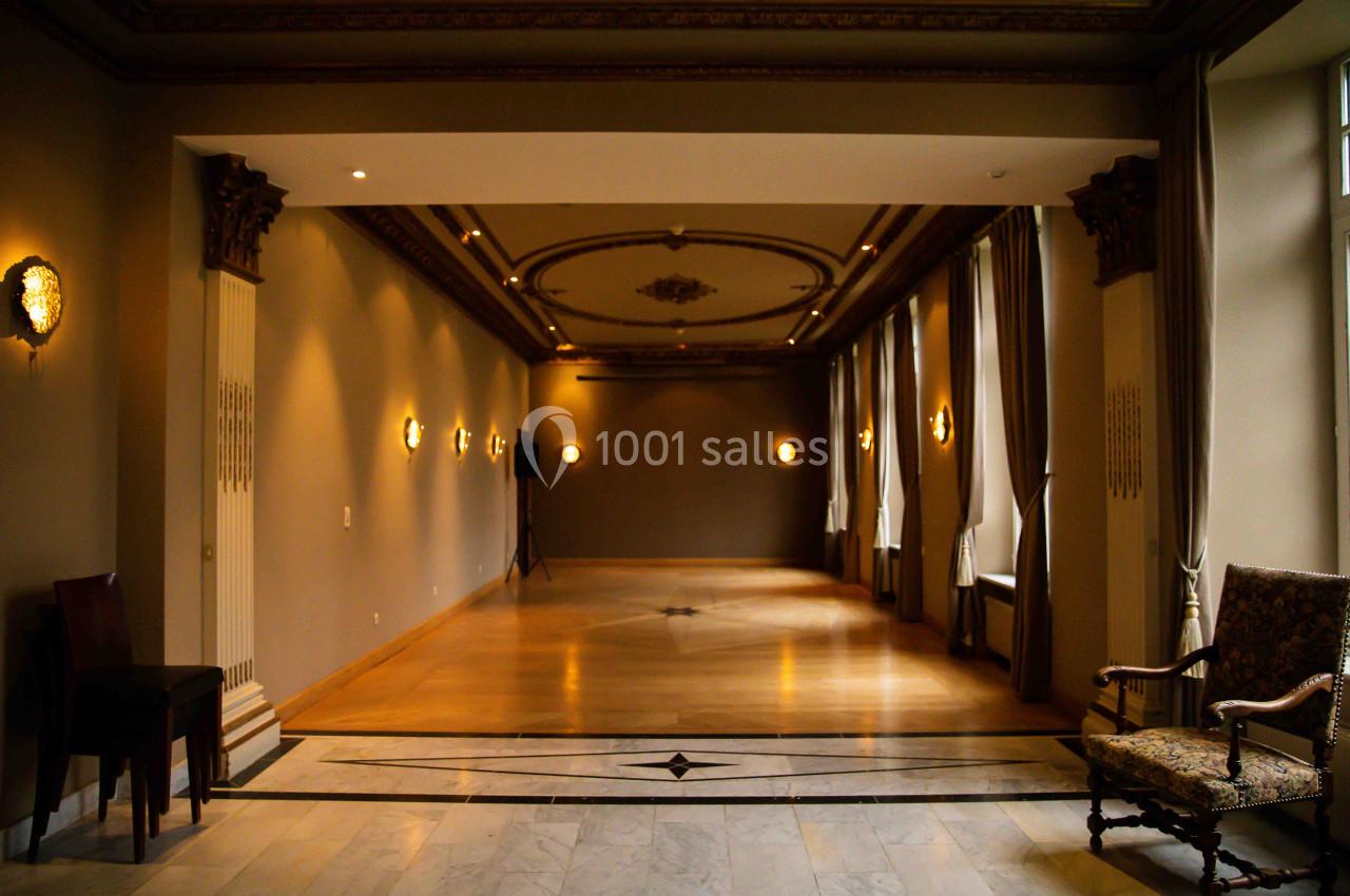 Salle élégante avec parquet, moulures au plafond, murs beige, appliques murales et grandes fenêtres avec rideaux.