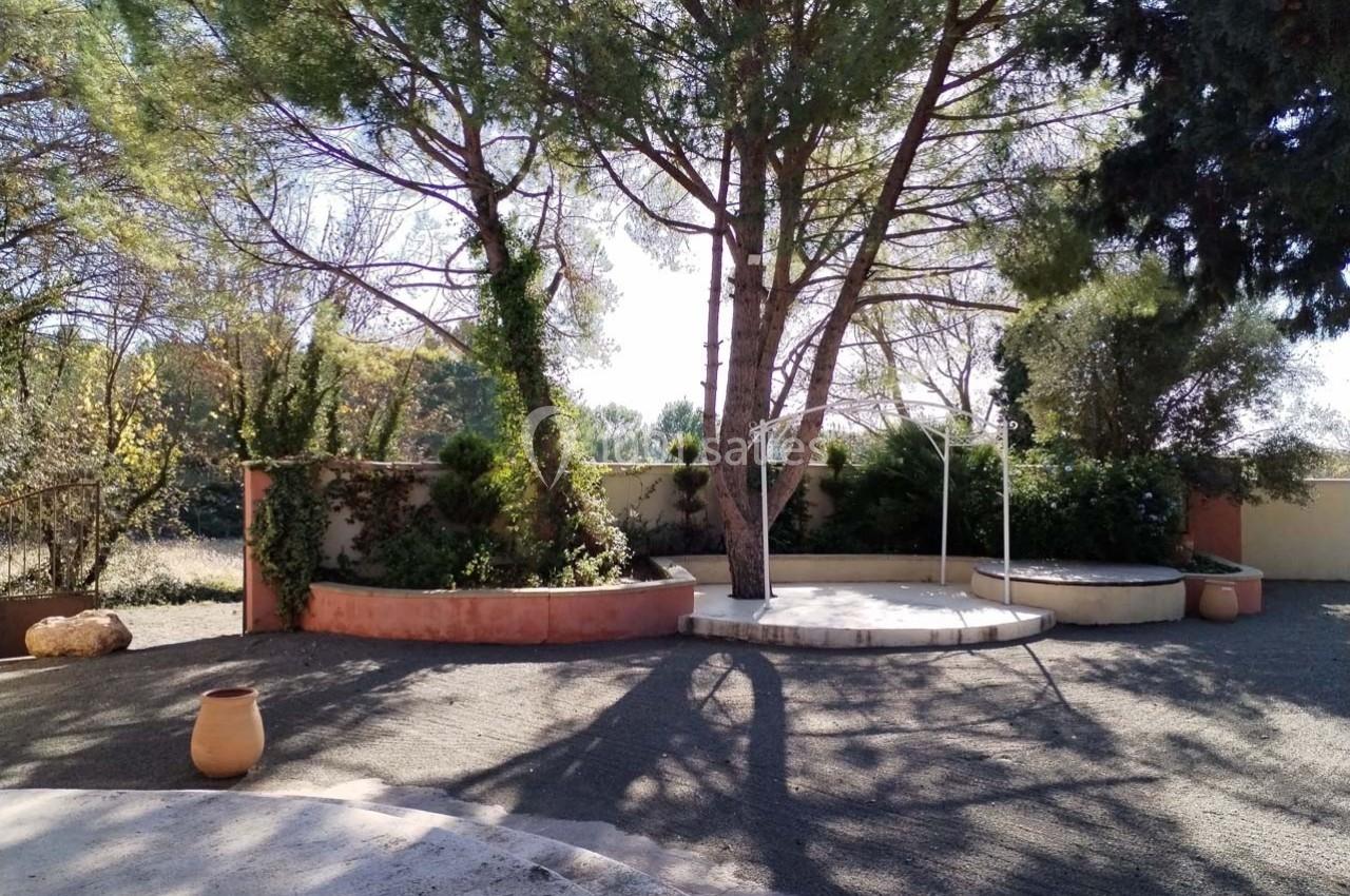 Cour extérieure avec arbres, muret en pierre, sol gravillonné et pot en terre cuite sous un ciel dégagé.