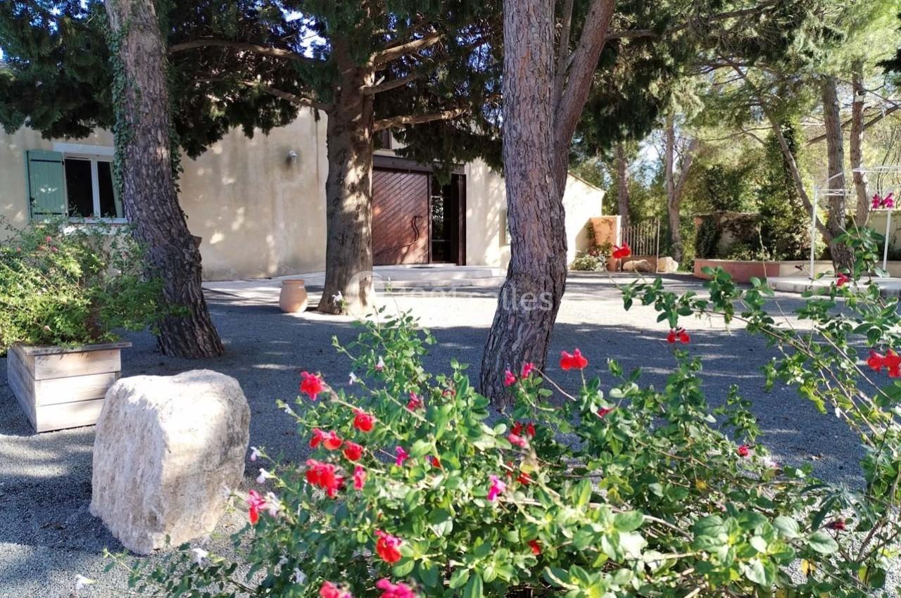 Jardin avec des arbustes fleuris rouges, arbres ombrageant une maison beige aux volets verts et porte en bois.
