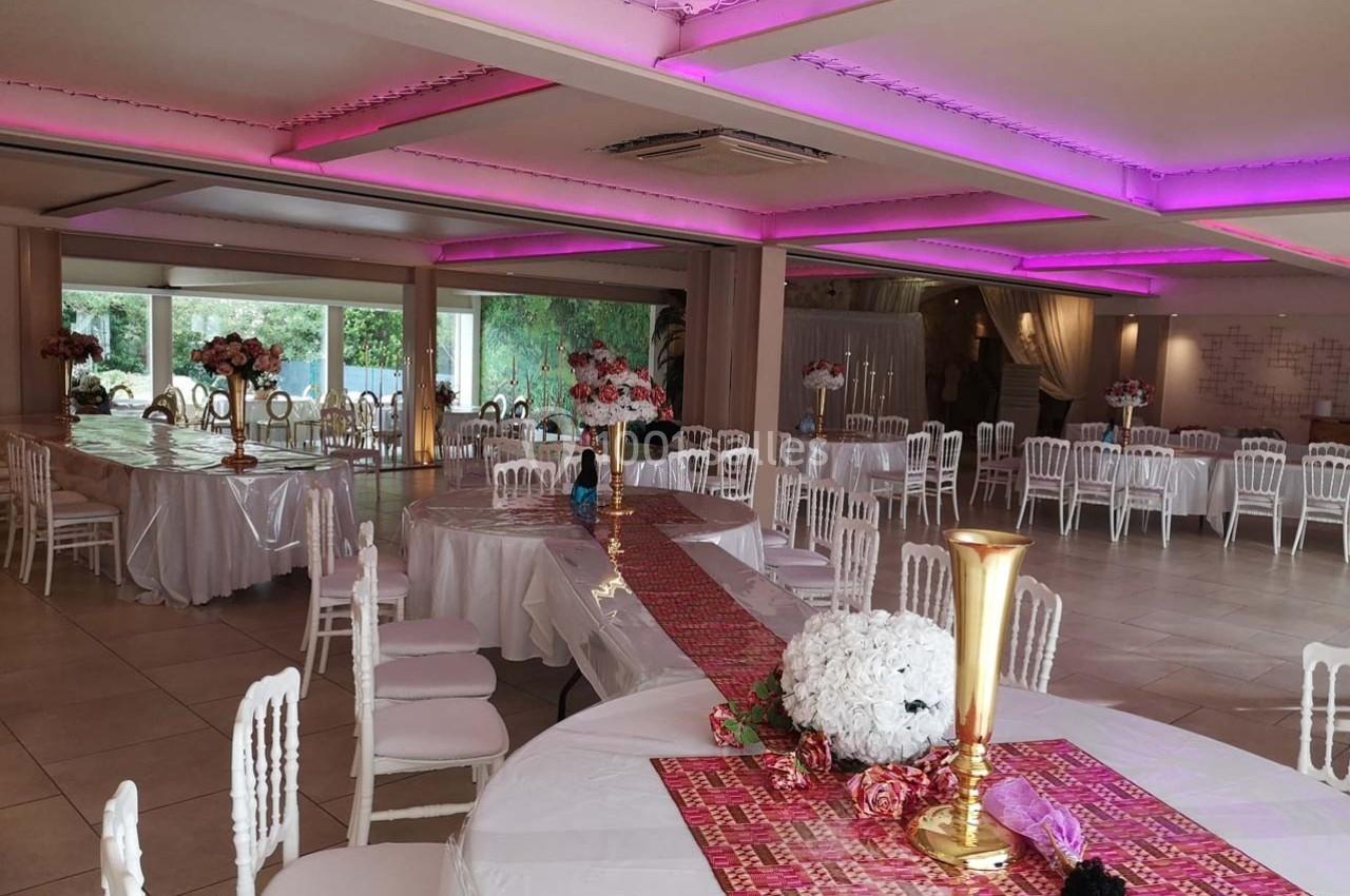 Salle de réception décorée avec des tables rondes, nappes blanches, centres de table fleuris et éclairage rose au plafond.