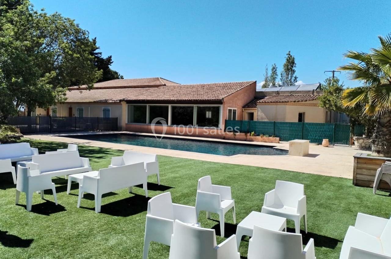 Espace extérieur avec piscine, pelouse artificielle, mobilier blanc et bâtiment en arrière-plan sous un ciel dégagé.