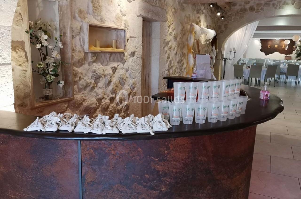 Comptoir en bois sombre avec des cadeaux et gobelets alignés, dans une salle en pierre décorée pour un événement.