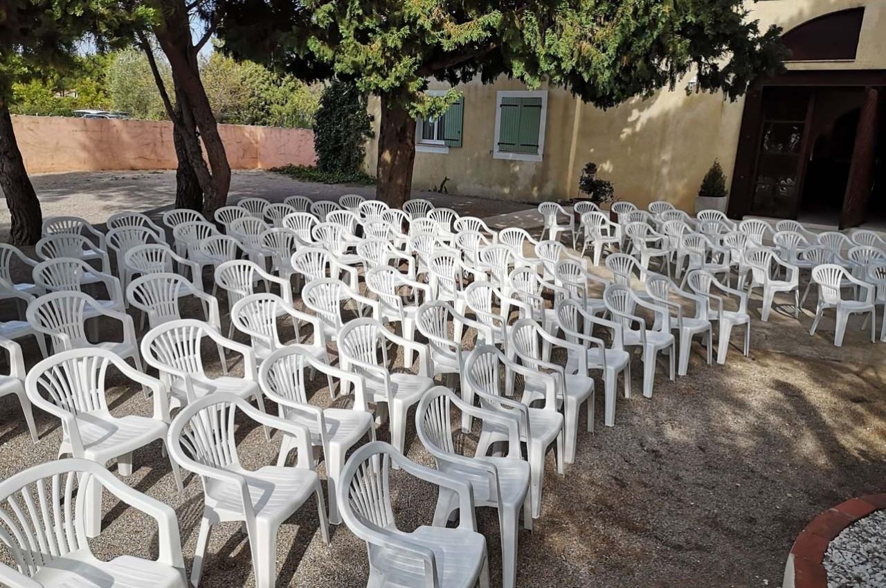 Rangées de chaises blanches en plastique disposées en extérieur sous des arbres, près d'un bâtiment beige.