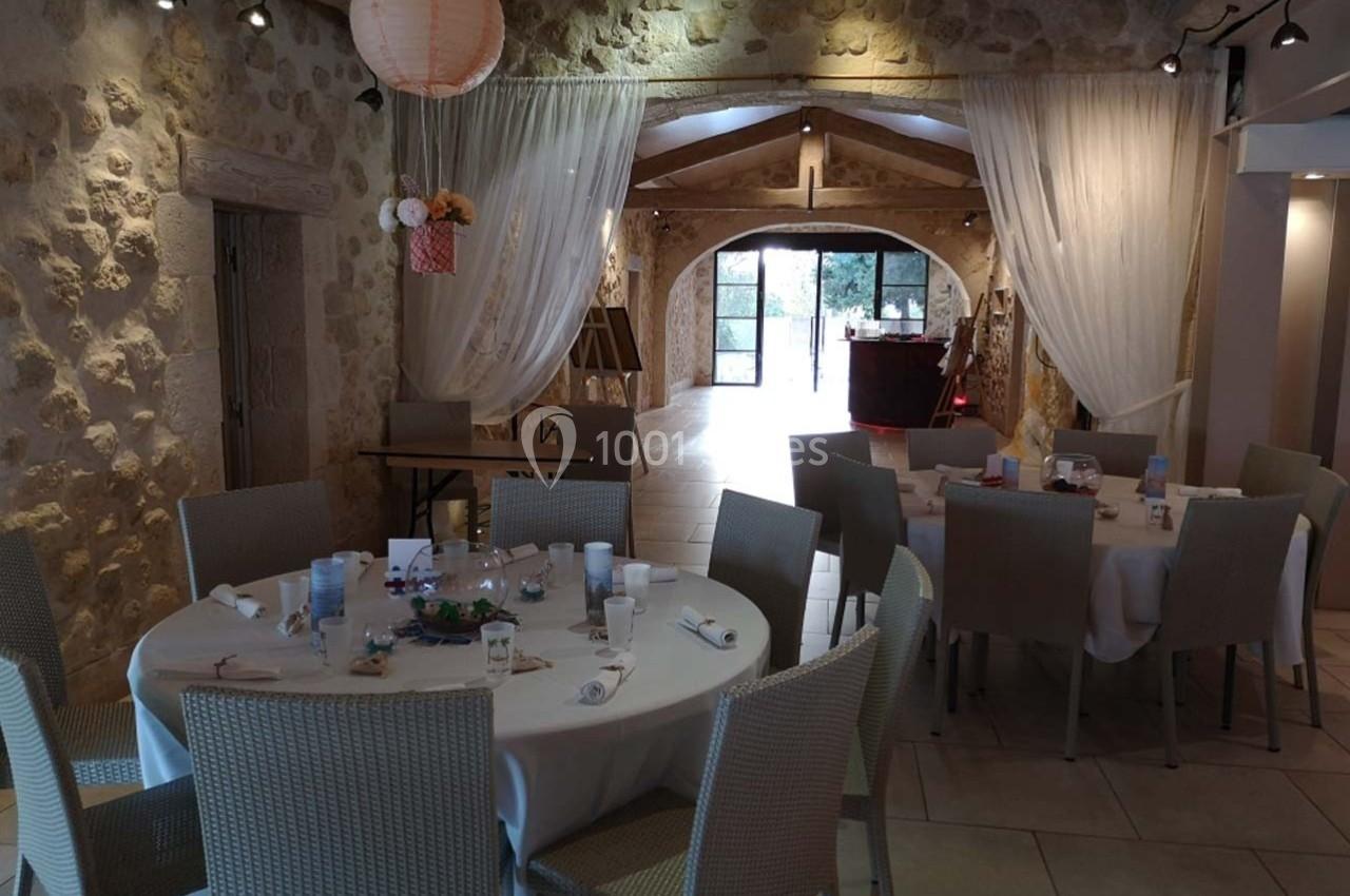 Salle de réception en pierre avec tables dressées, chaises en osier clair et rideaux blancs encadrant une entrée lumineuse.