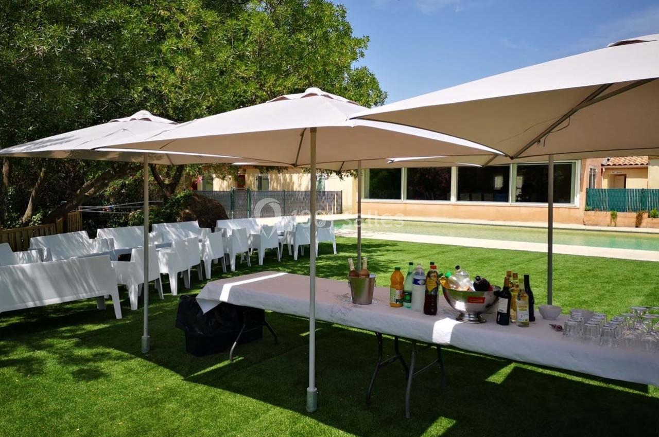 Tables et chaises blanches disposées sous des parasols dans un jardin, avec des boissons et verres sur une table.
