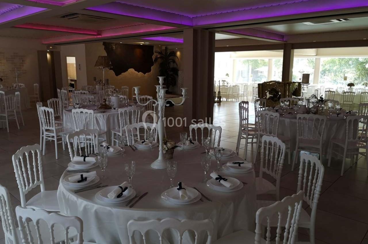 Salle de réception décorée avec des tables rondes, nappes blanches, chaises élégantes et éclairage tamisé.