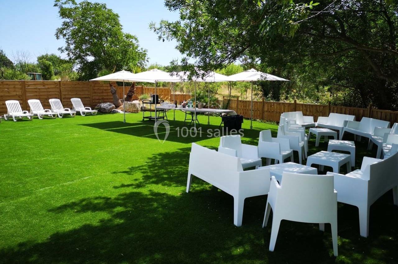 Espace extérieur avec pelouse artificielle, chaises blanches, tables et parasols, entouré de clôtures en bois et d'arbres.