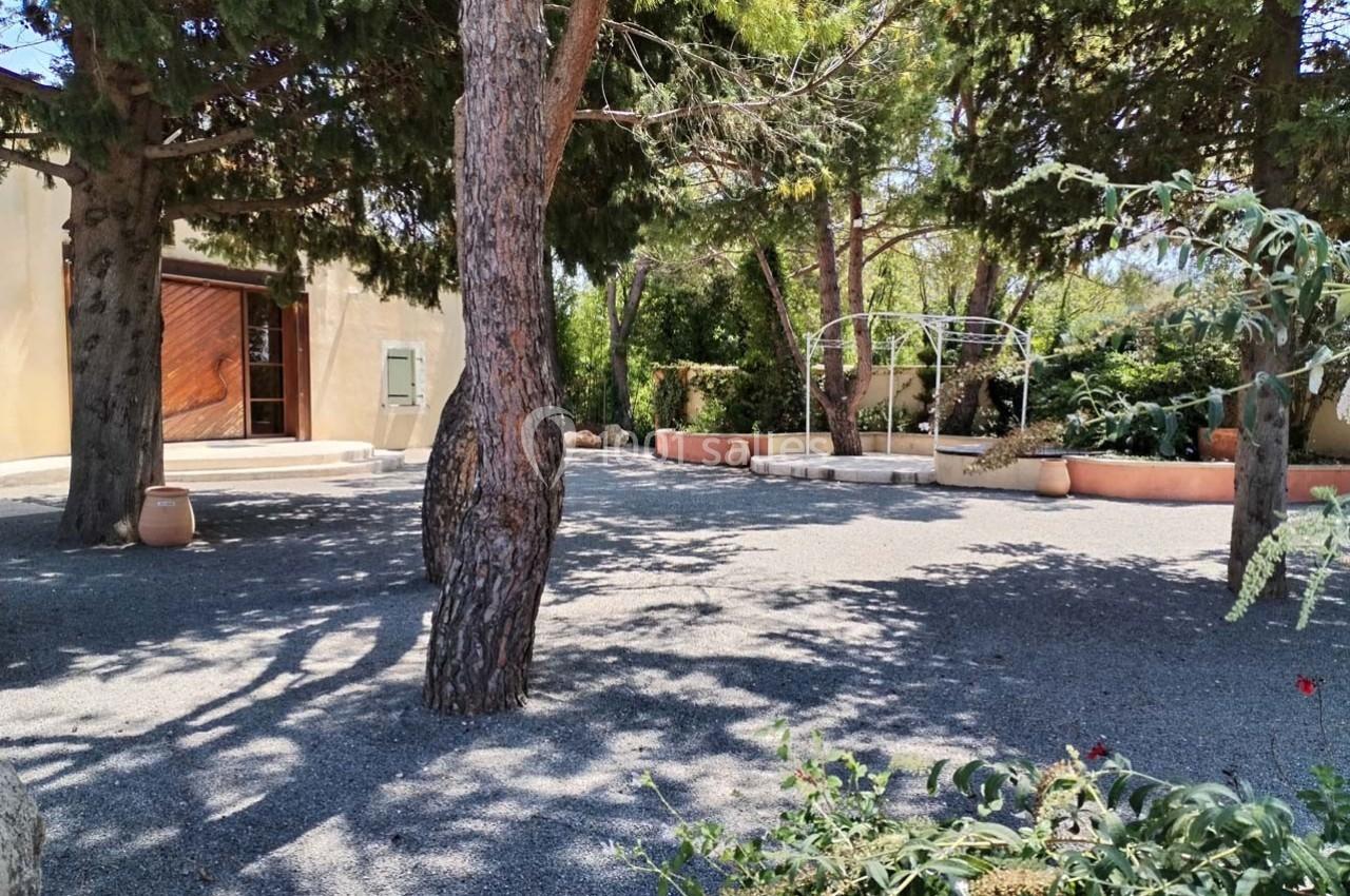 Cour extérieure ombragée avec des arbres, sol en gravier, pots en terre cuite et structure métallique en arrière-plan.