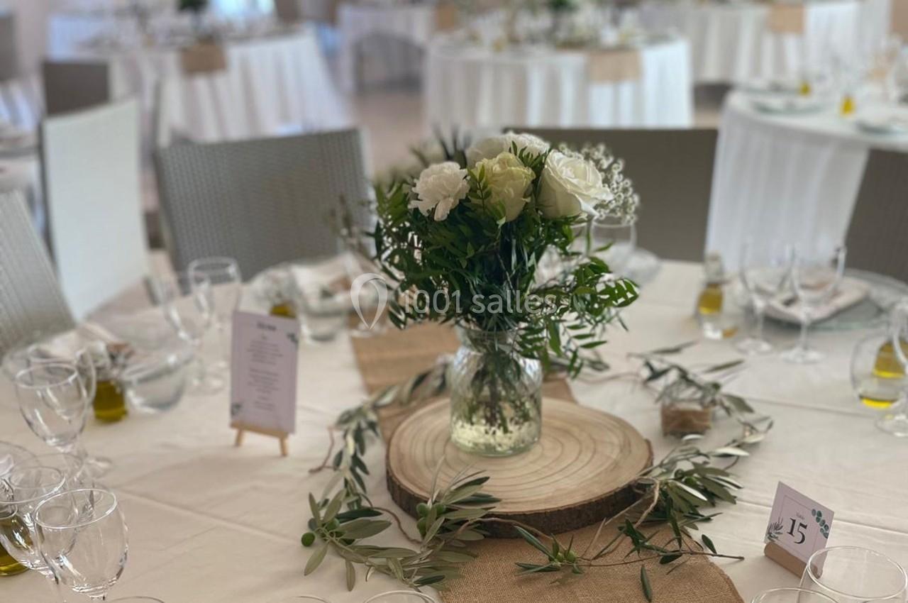 Centre de table avec vase de fleurs blanches et feuillage, posé sur une rondelle de bois, entouré de branches d’olivier.