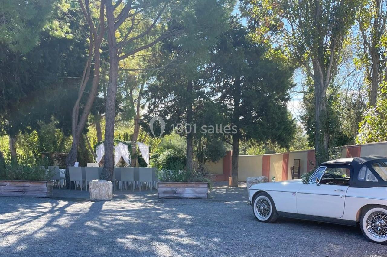 Voiture ancienne blanche garée sur un parking ombragé près d'une table dressée sous des arbres.