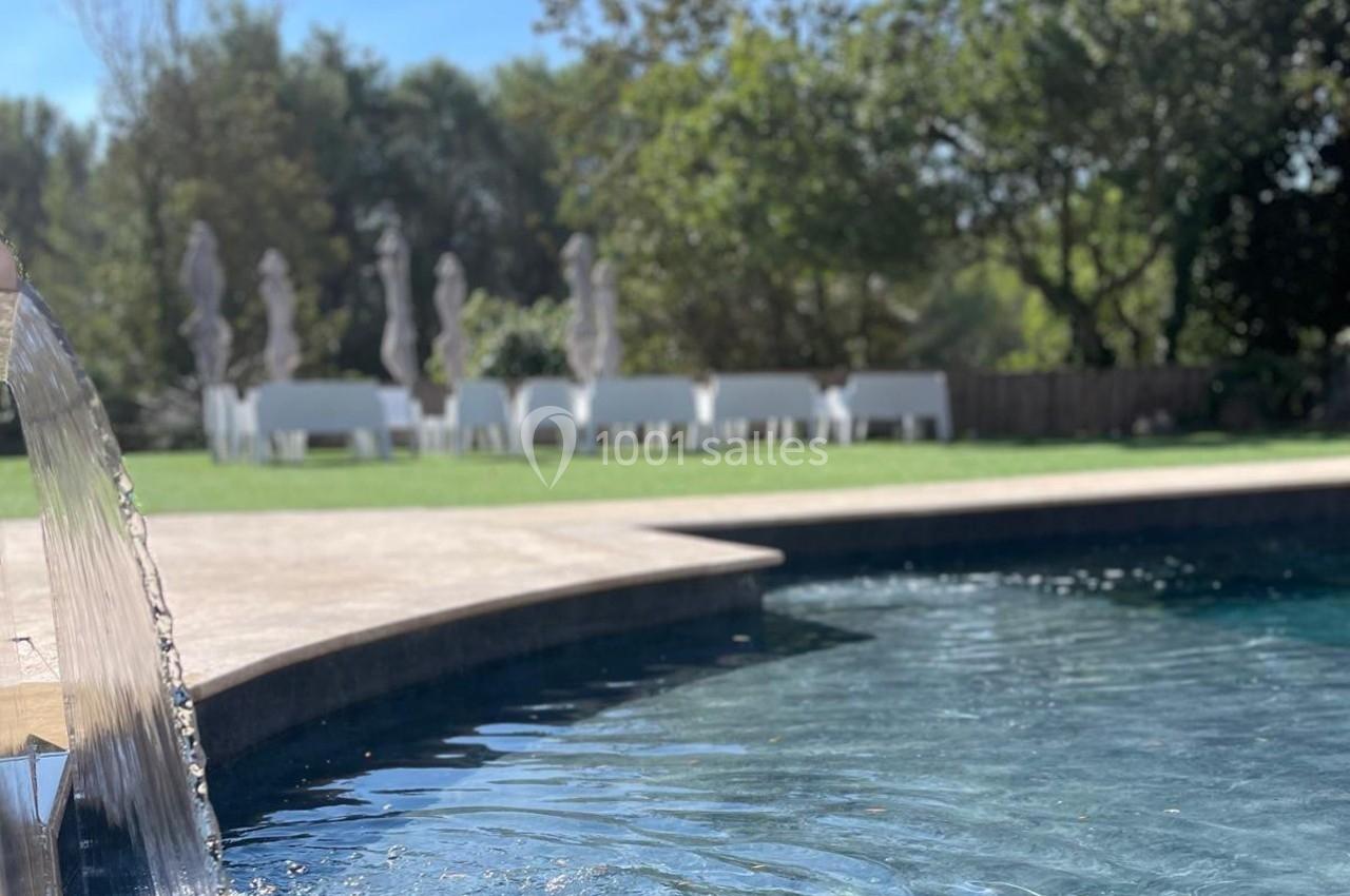 Eau d'une piscine avec une cascade au premier plan, pelouse et tables blanches sous des parasols en arrière-plan.