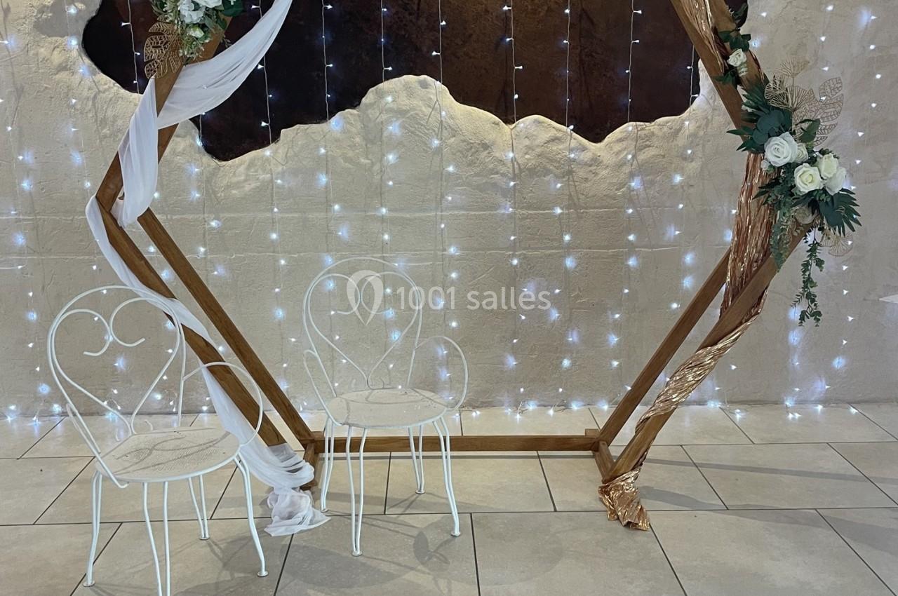 Deux chaises blanches en métal devant une arche en bois décorée de fleurs et de voilages, avec un fond lumineux.