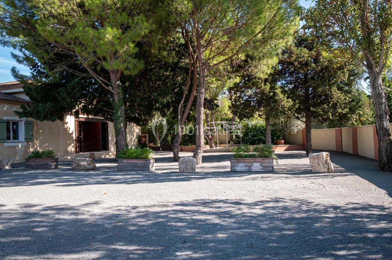 Cour extérieure ombragée avec des arbres, des jardinières en bois, un sol en gravier et un bâtiment en arrière-plan.