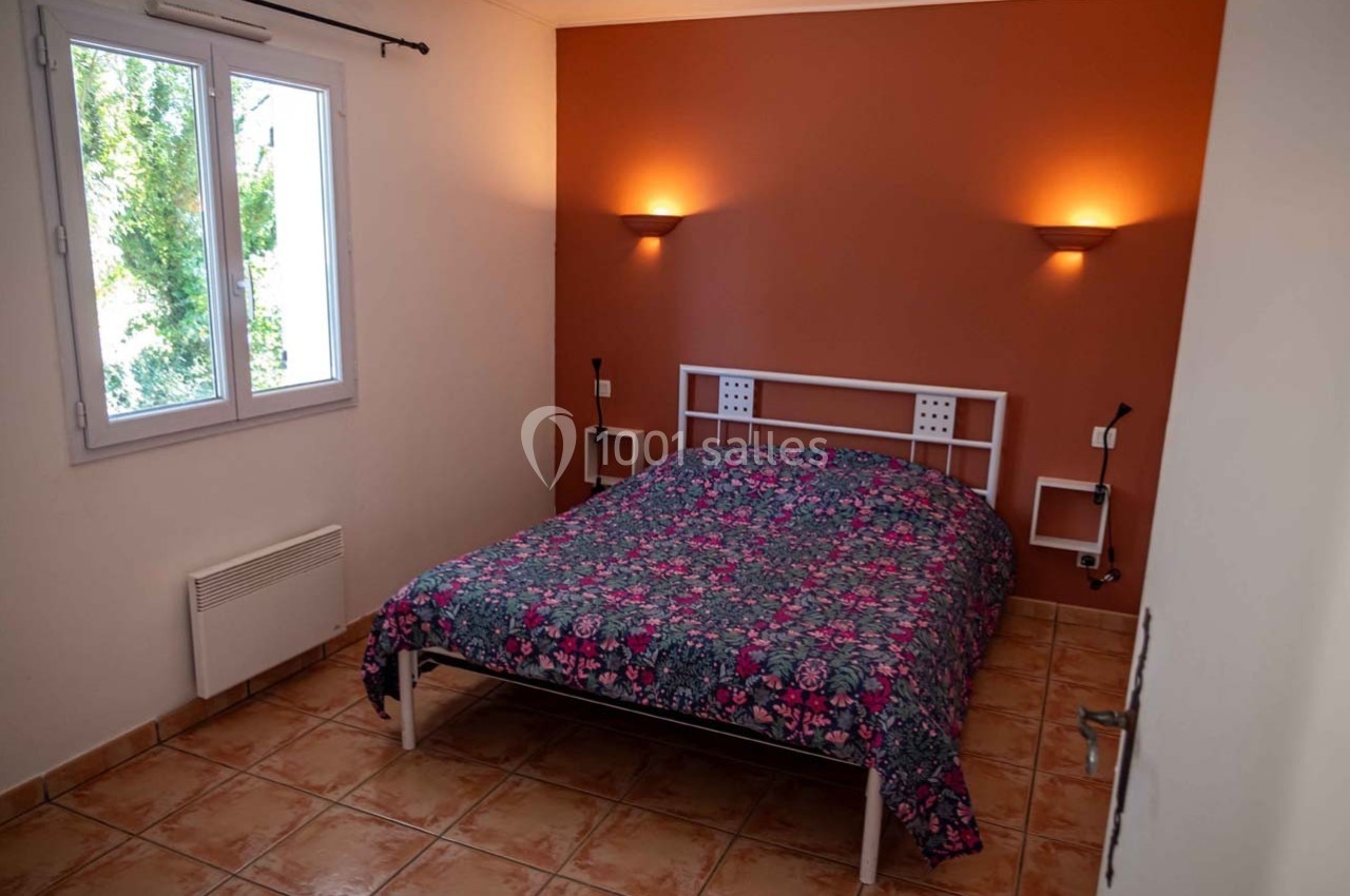 Chambre avec un lit double, mur orange, carrelage au sol, fenêtre à gauche et appliques murales allumées.