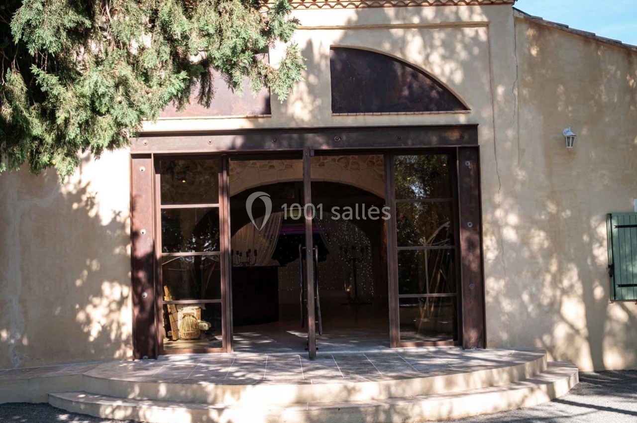 Entrée d'un bâtiment avec une porte vitrée ouverte, entourée d'arbres projetant des ombres sur la façade beige.