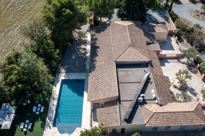 Vue aérienne d'une maison avec toit en tuiles, piscine rectangulaire et jardin arboré dans un cadre rural.