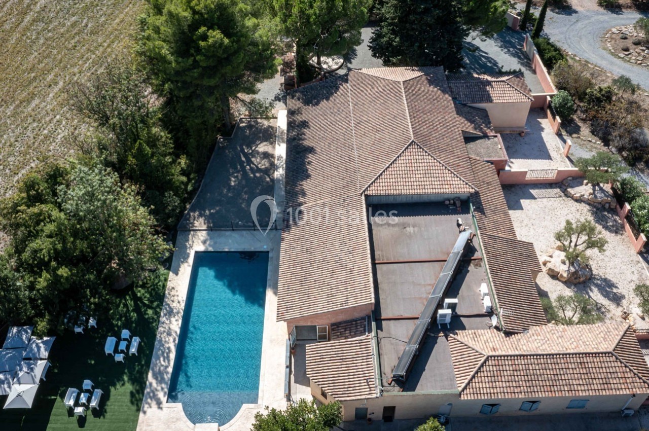 Vue aérienne d'une maison avec toit en tuiles, piscine rectangulaire et jardin arboré dans un cadre rural.