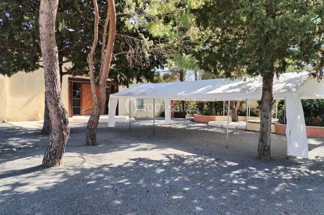 Tente blanche installée dans une cour extérieure ombragée par des arbres, avec un bâtiment en arrière-plan.