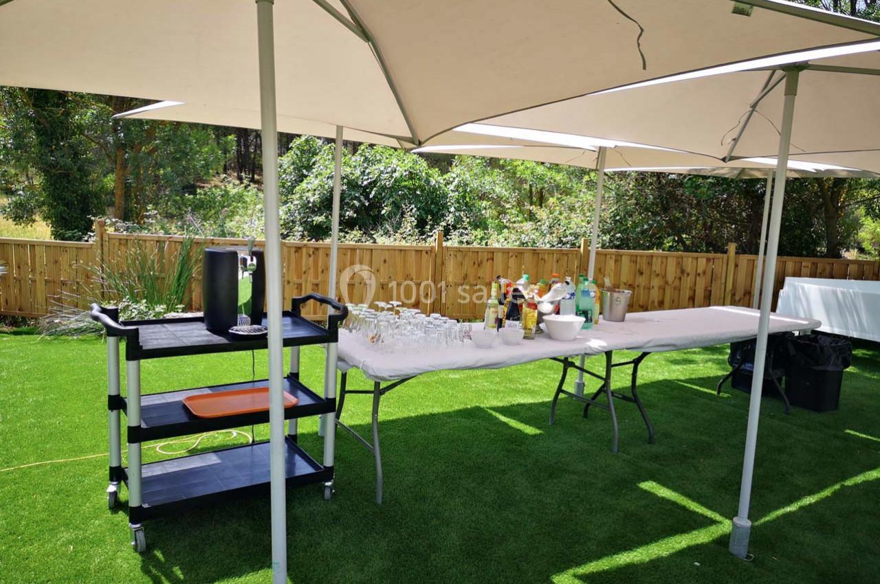 Table de buffet en extérieur avec boissons, verres et parasols, installée sur une pelouse près d'une clôture en bois.