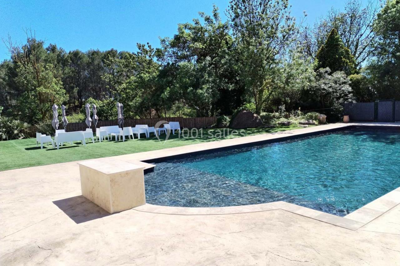 Piscine extérieure entourée de verdure avec des chaises blanches et des parasols sur une pelouse.