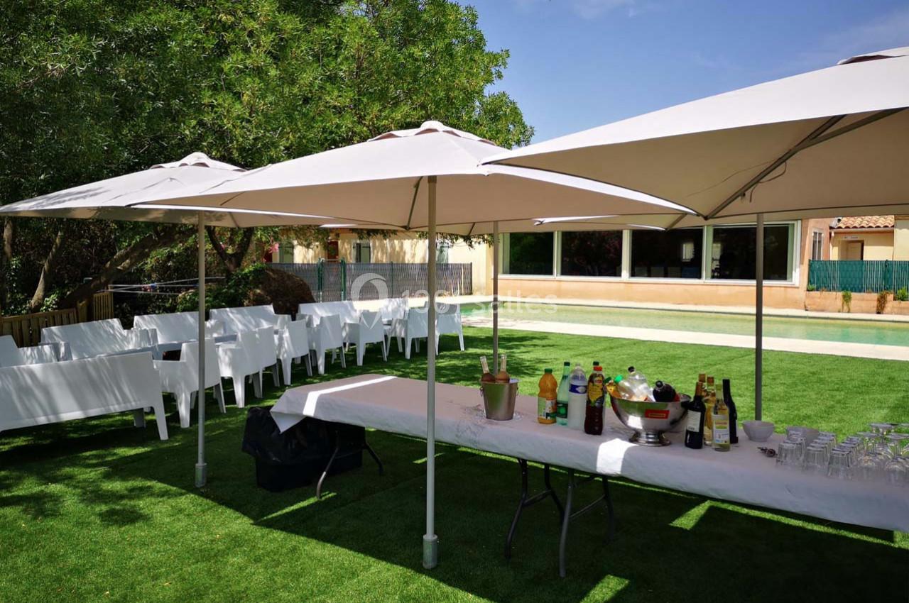 Table dressée avec boissons et verres sous des parasols blancs, entourée de chaises blanches sur une pelouse.