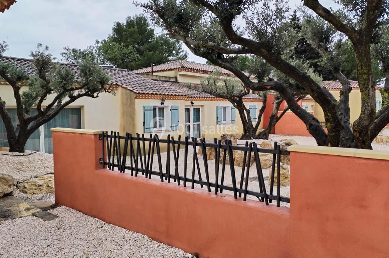 Maisons de plain-pied avec façades colorées, entourées d'oliviers et de graviers, derrière une clôture en métal noir.