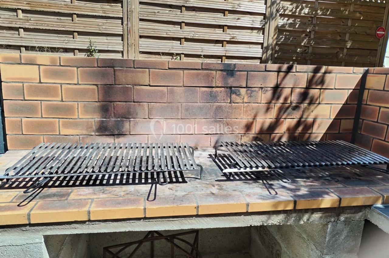Barbecue en briques avec deux grilles métalliques, situé dans un espace extérieur avec une clôture en bois en arrière-plan.