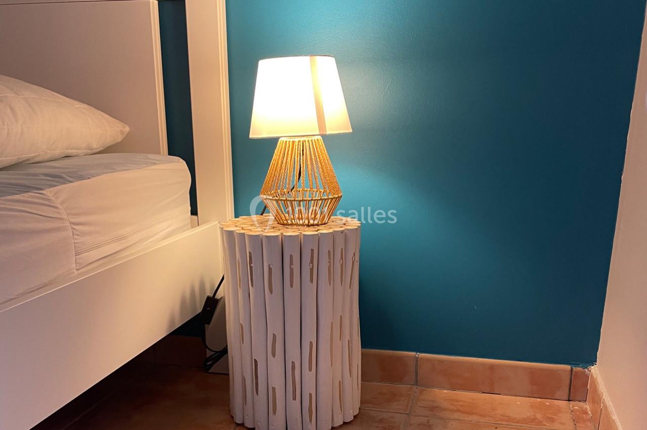 Lampe allumée sur une table de chevet en bois blanc, près d'un lit avec mur bleu en arrière-plan.