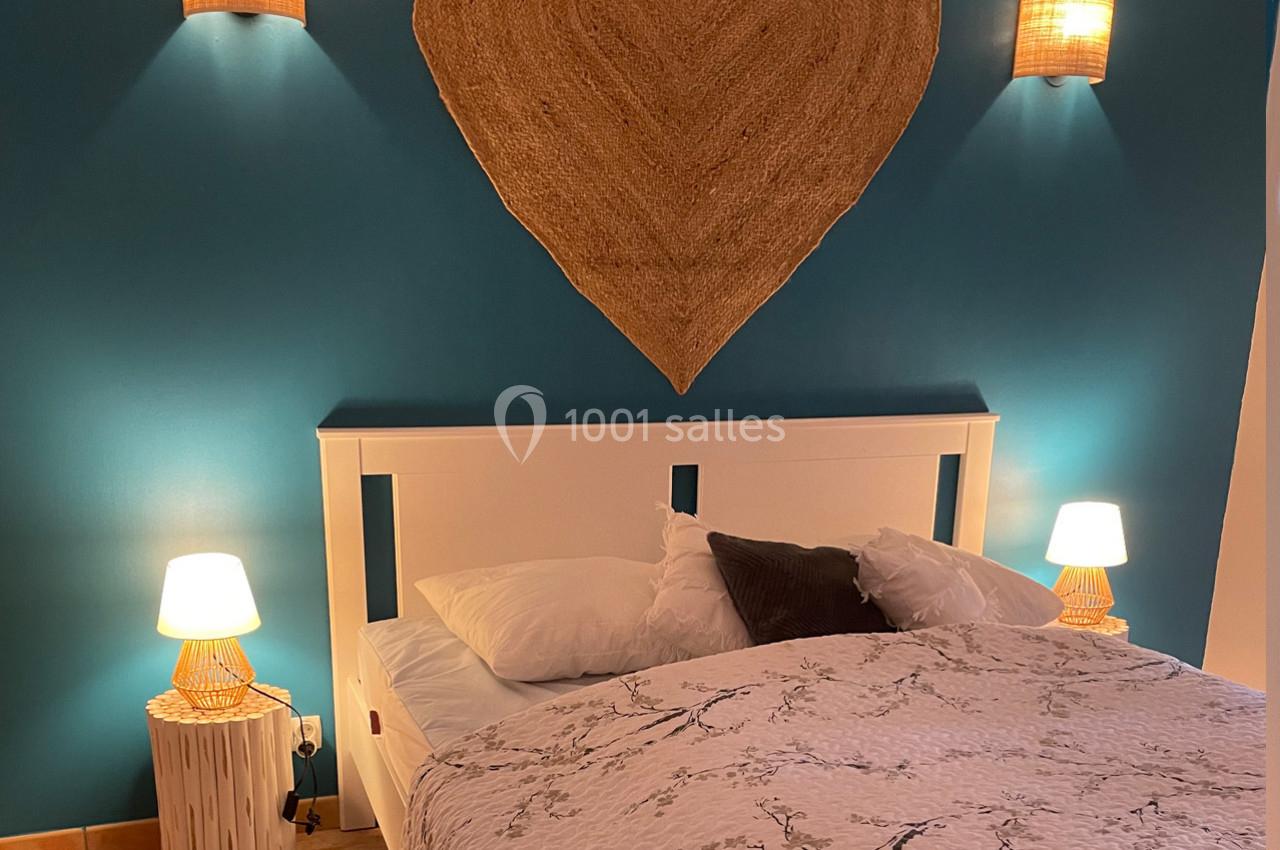 Lit double avec draps à motifs floraux, encadré par deux lampes de chevet et un mur bleu décoré d'un cœur en rotin.