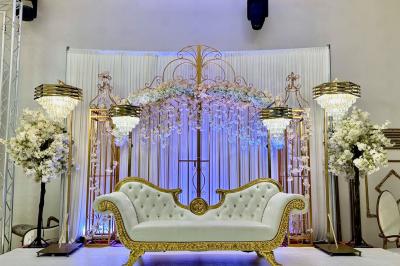 Salle de réception décorée avec des tables élégantes, des bouquets de fleurs et une estrade ornée d'un canapé doré.
