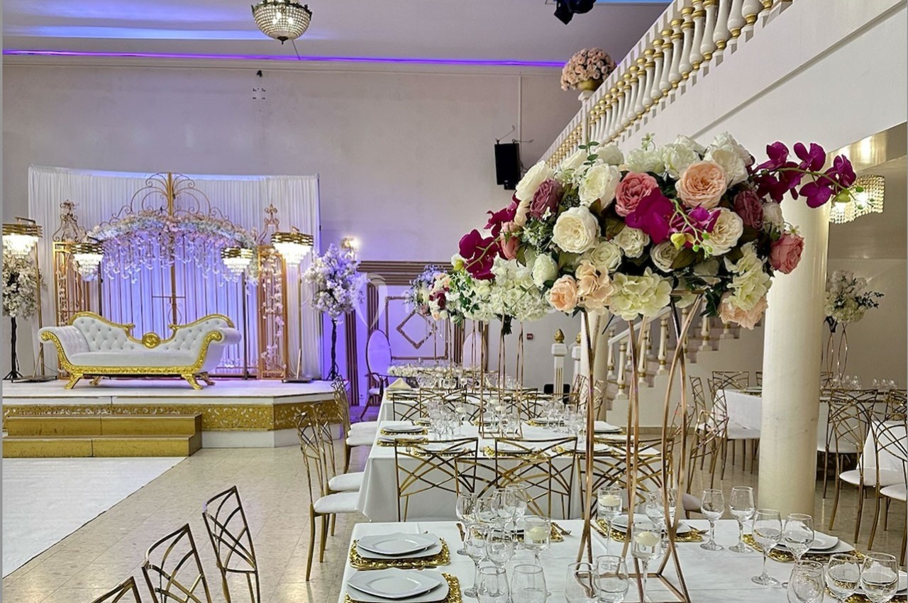Salle de réception décorée avec des tables élégantes, des bouquets de fleurs et une estrade ornée d'un canapé doré.
