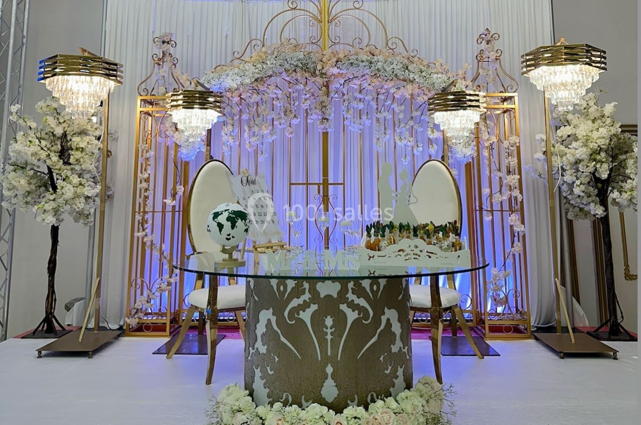 Décoration de table de mariage avec chaises élégantes, fleurs blanches, lustres et ornements dorés sur fond lumineux.
