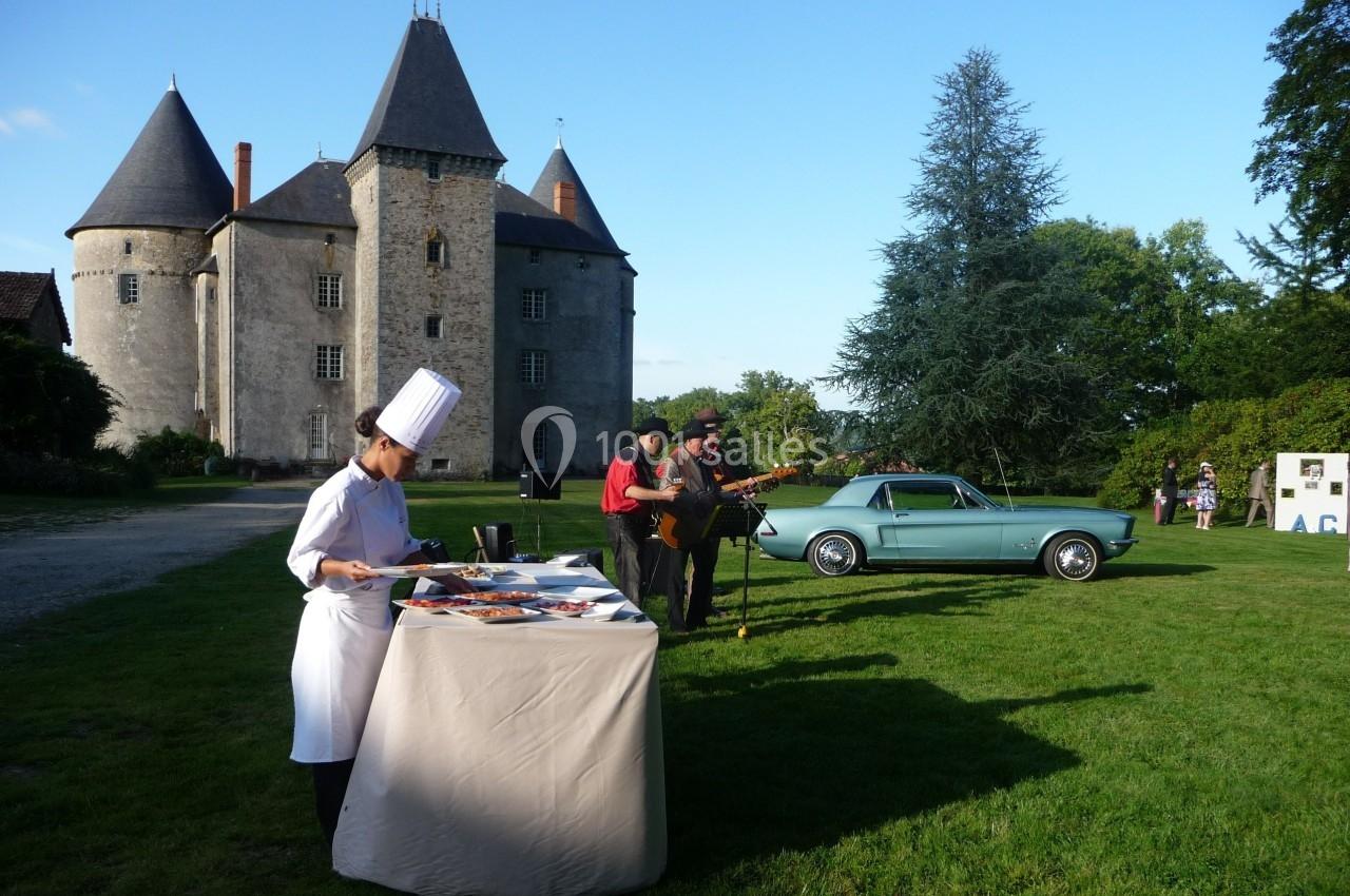 Chef en tenue blanche préparant des plats sur une table en extérieur, devant un château, avec musiciens et voiture ancienne.