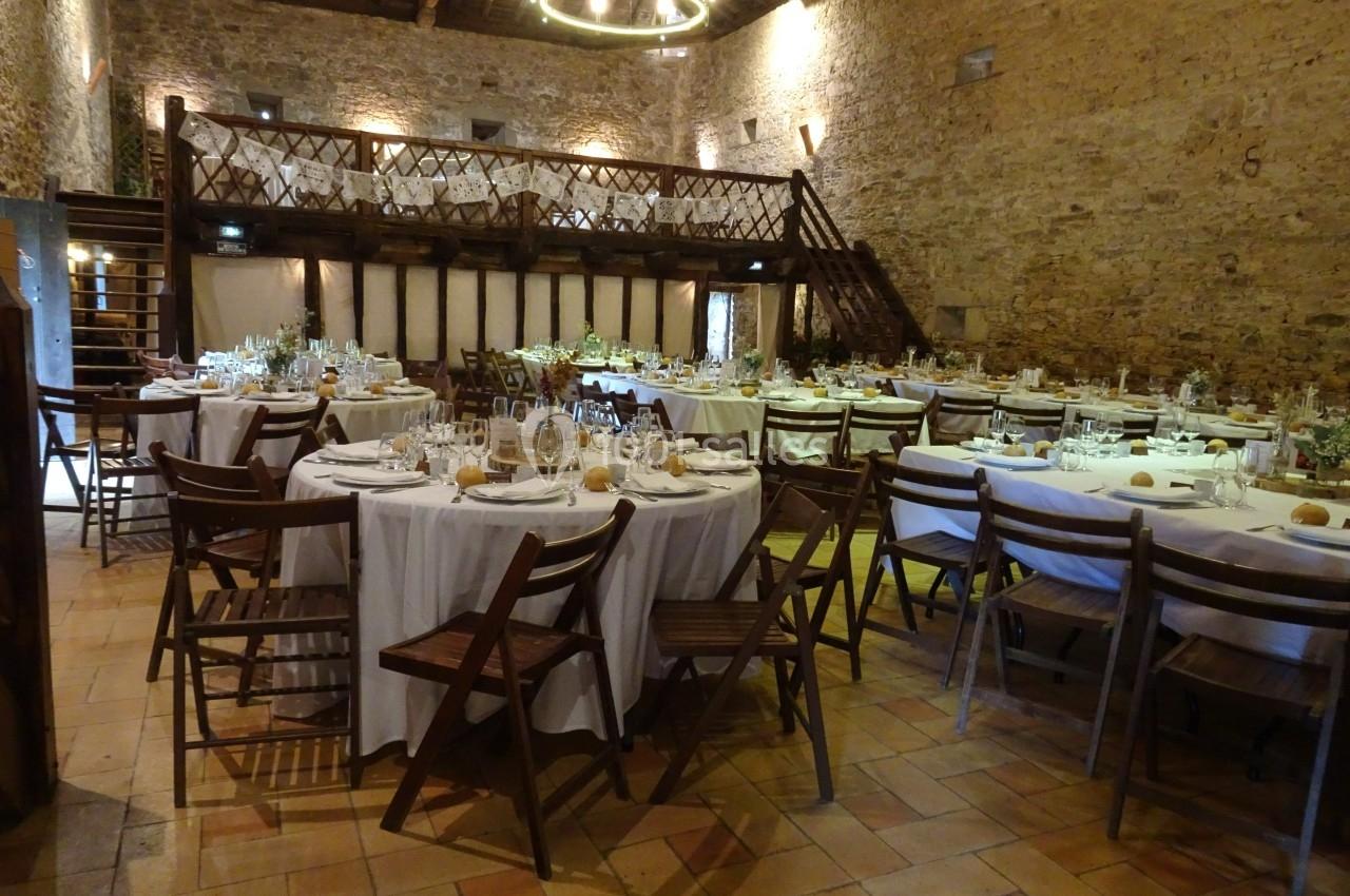 Salle de réception rustique avec tables rondes dressées, chaises en bois et mezzanine en pierre apparente.