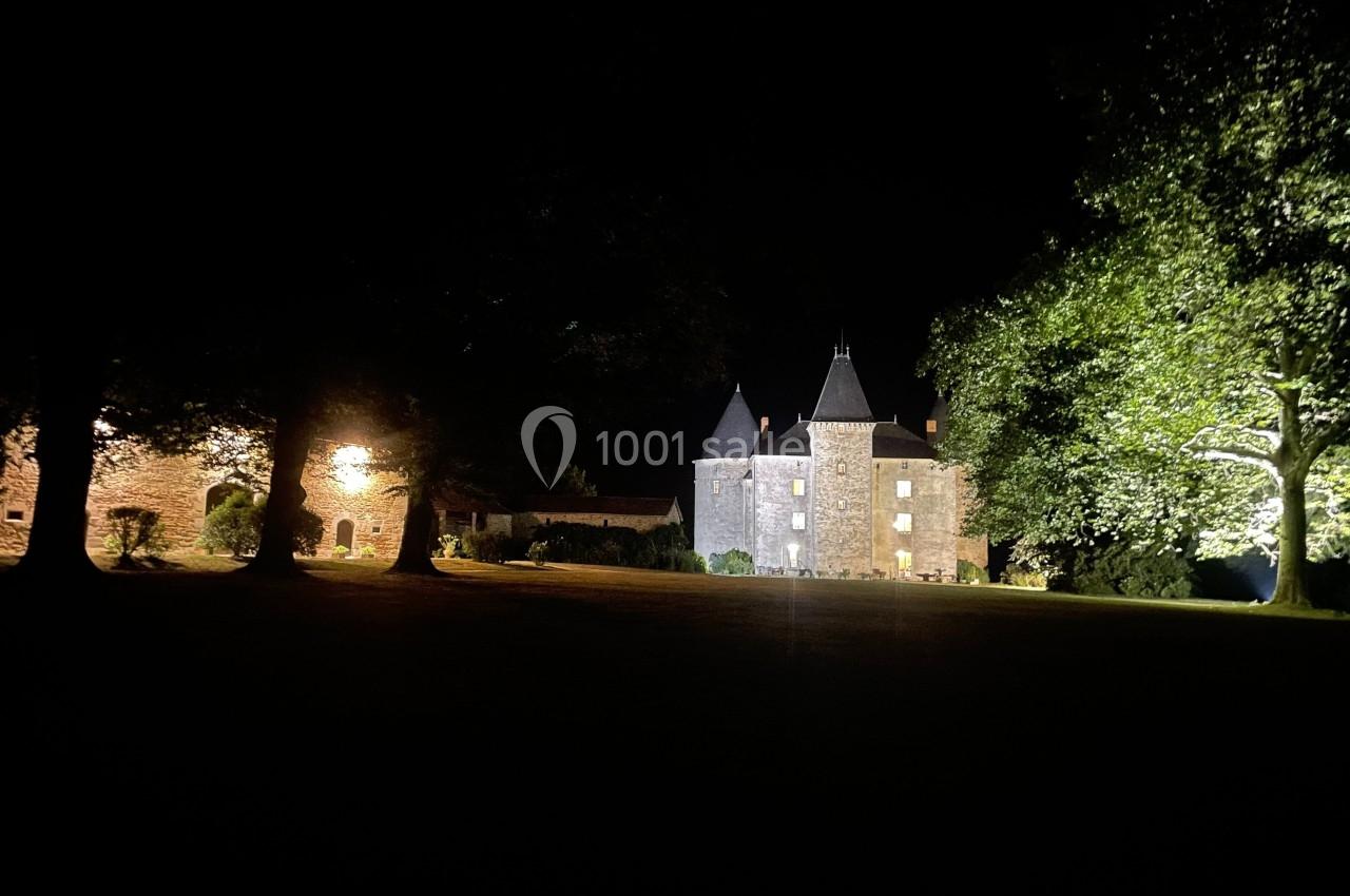 Château éclairé de nuit entouré d'arbres, avec une façade en pierre et des tours coniques.