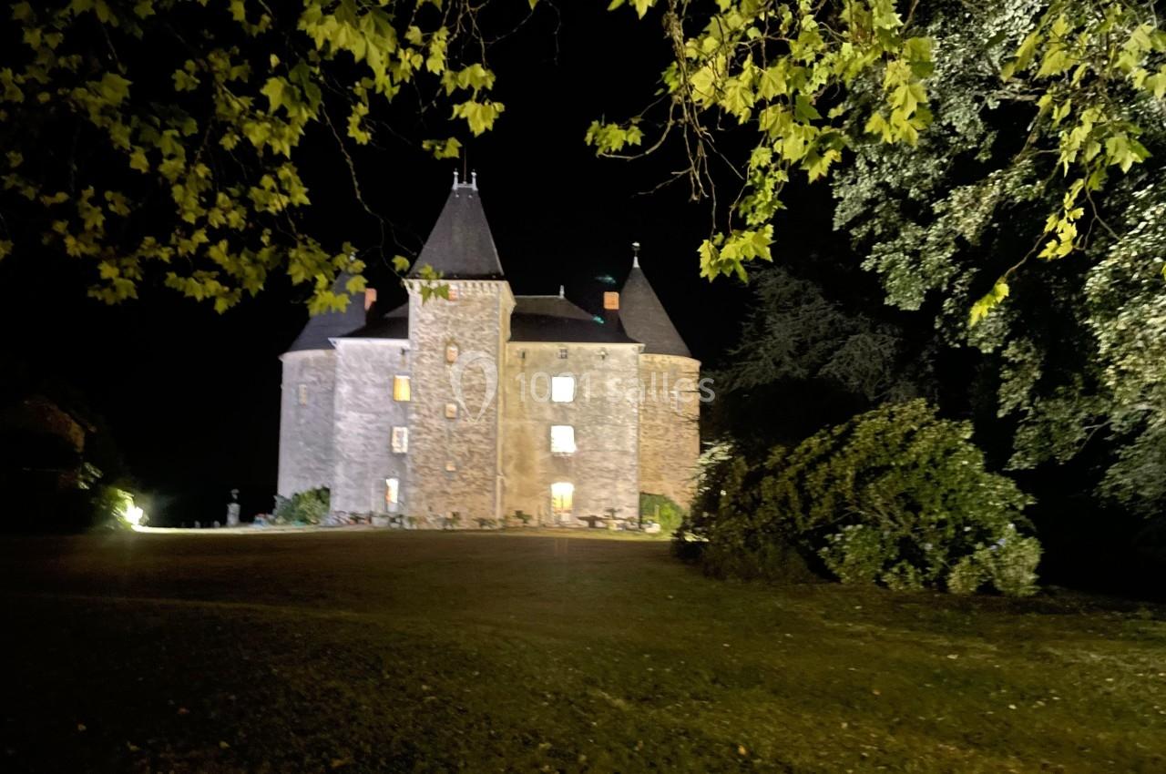 Château en pierre éclairé de nuit, entouré d'arbres et d'une pelouse sombre.