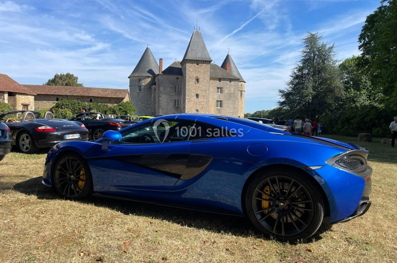 Voiture de sport bleue garée sur une pelouse devant un château ancien sous un ciel dégagé.